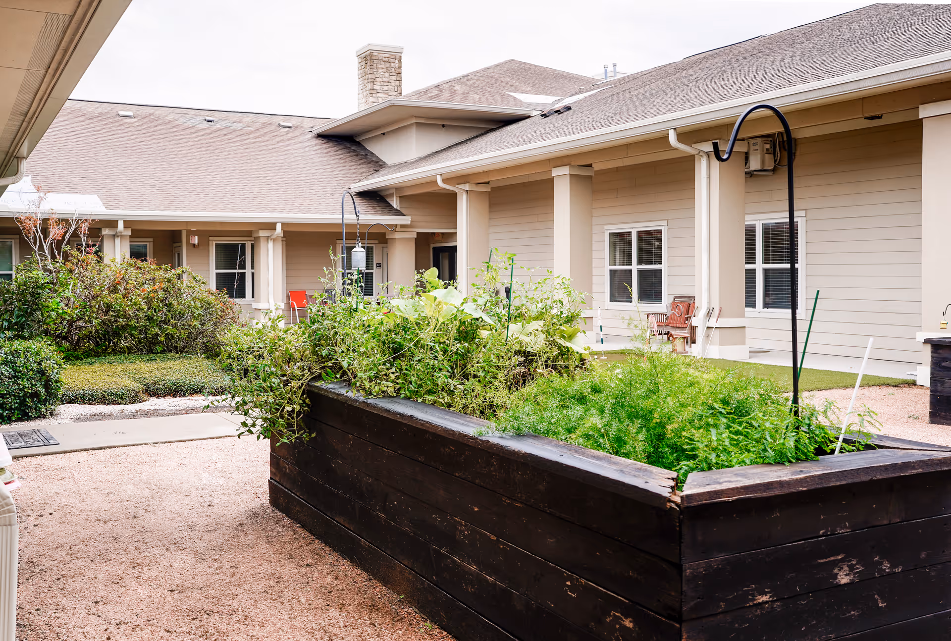 Raised wooden planter filled with greenery in the courtyard of a single-story assisted living building with columns, windows and outdoor seating.
