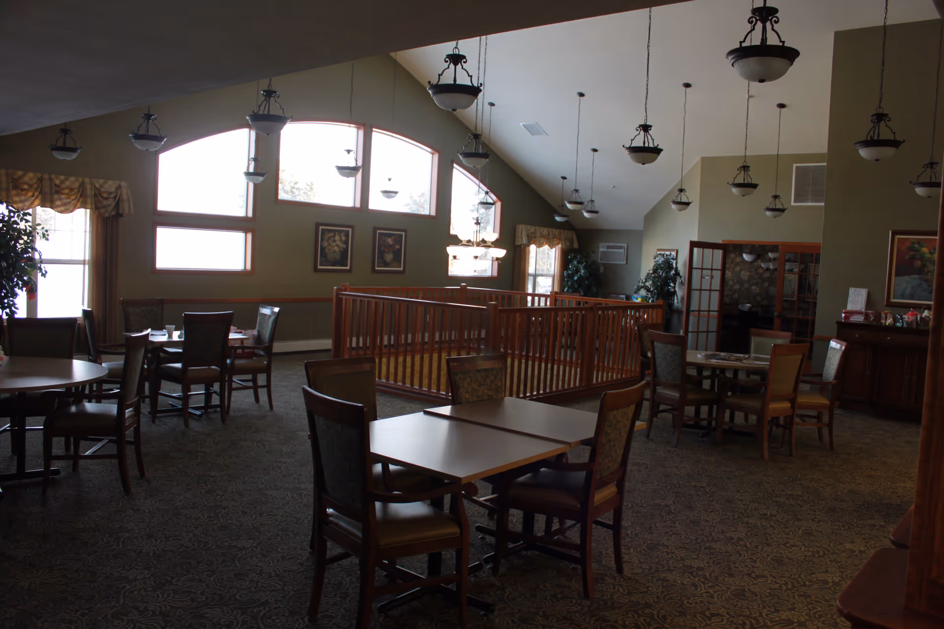 Interior view of a senior living facility dining area with multiple tables and chairs arranged on a patterned carpet. Large windows with arched tops allow natural light to enter the room. Several hanging light fixtures are suspended from the high ceiling. The walls are decorated with framed floral artwork and there are plants placed near the windows and walls. A wooden railing encloses a small area in the center of the room.