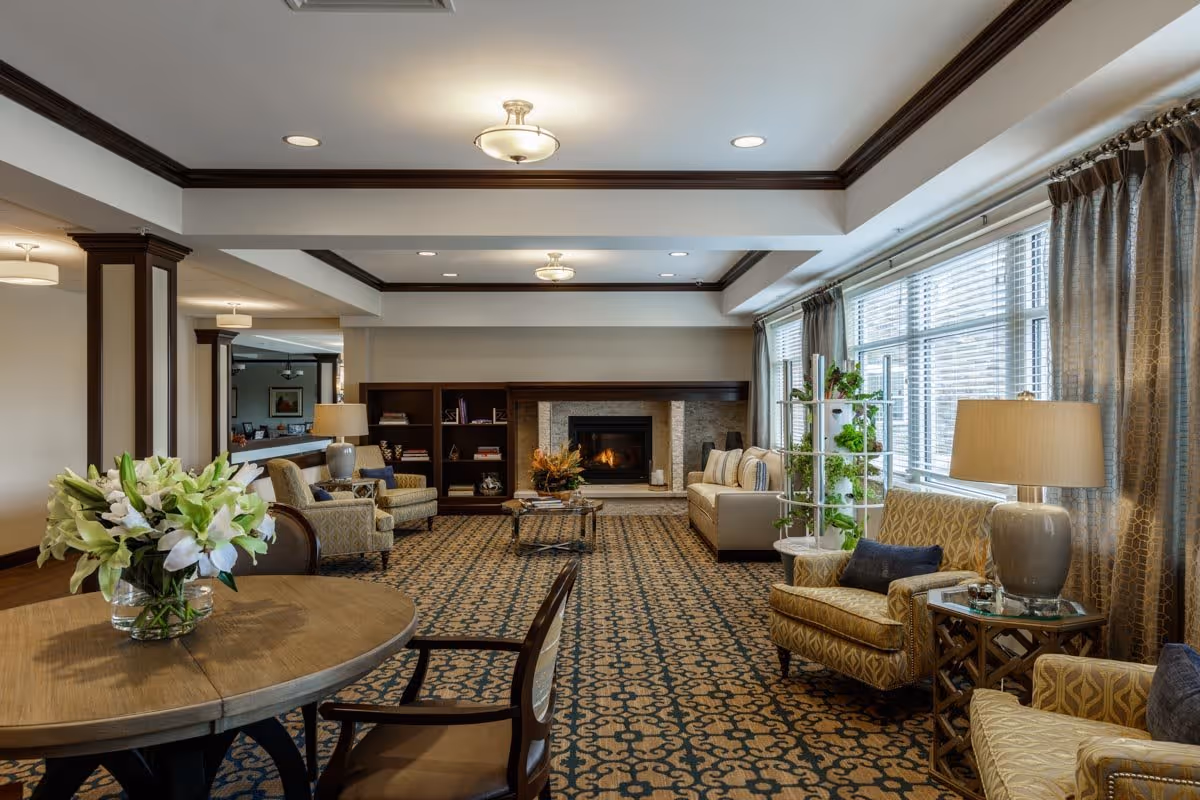 A cozy living room area in Clarendale of Algonquin featuring a patterned carpet, a round wooden table with a vase of white lilies, several upholstered armchairs and sofas, a fireplace with a stone surround, built-in bookshelves, large windows with curtains, and warm lighting from ceiling fixtures and table lamps.