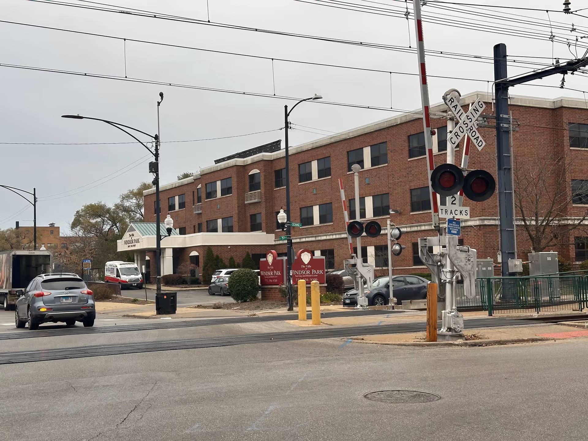 Exterior view of a three-story brick building with a sign that reads 'The Villa Windsor Park Nursing and Rehabilitation' near a railroad crossing with lowered gates and warning lights. Several cars and a delivery truck are visible on the street in front of the building under an overcast sky.