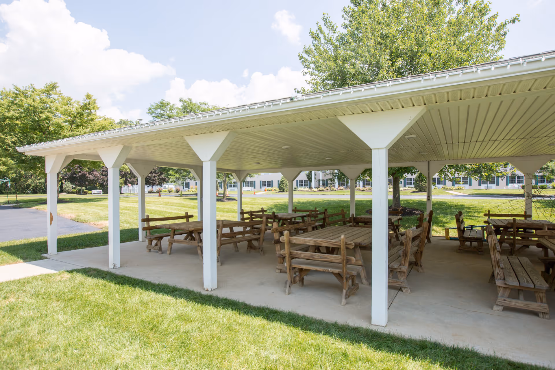 Covered outdoor pavilion with multiple wooden picnic tables and benches on a concrete floor, surrounded by green grass and trees, with a building visible in the background under a partly cloudy sky.