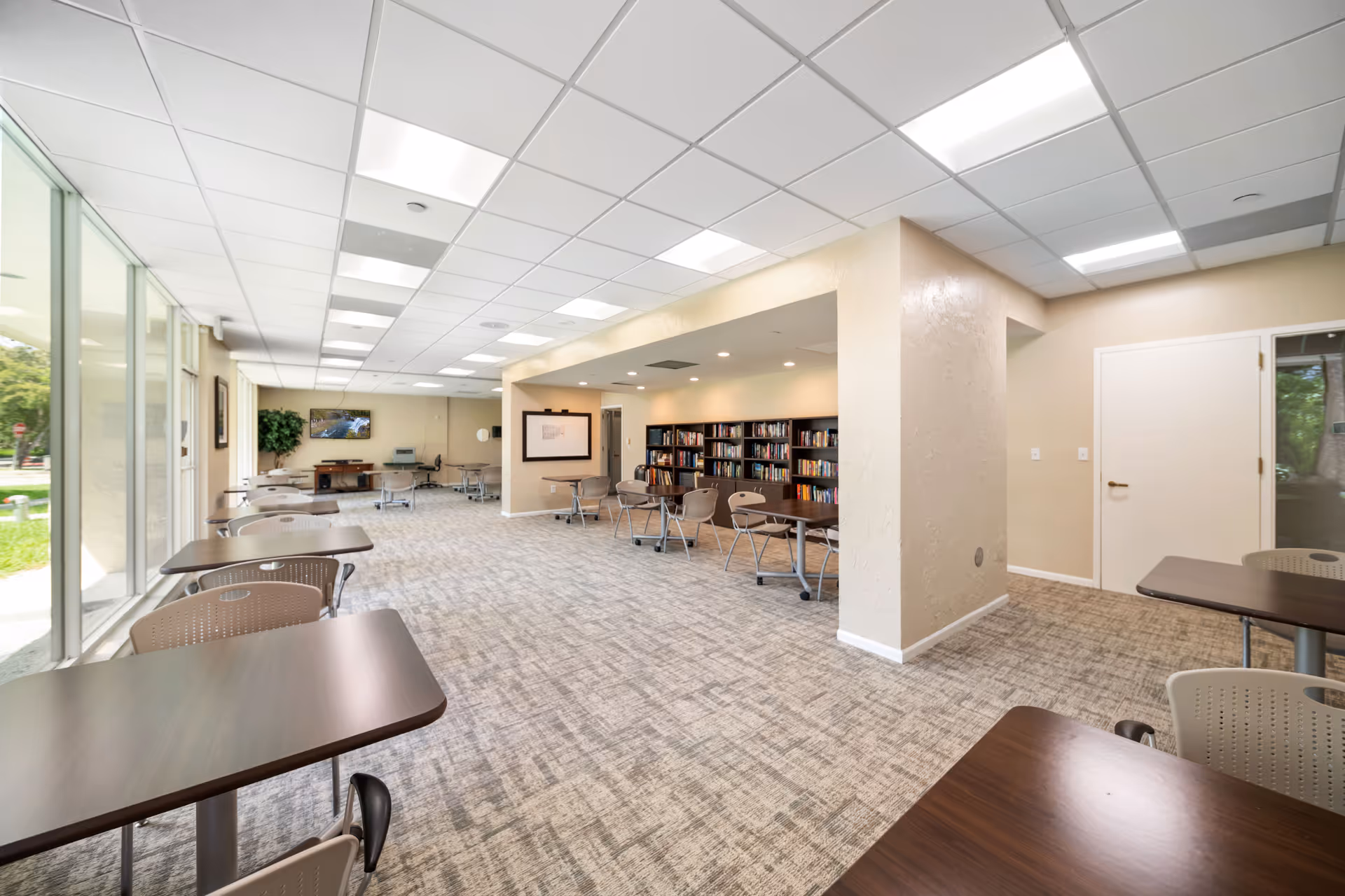 Bright communal activity room with tables and chairs, bookshelves, and large windows along one side.