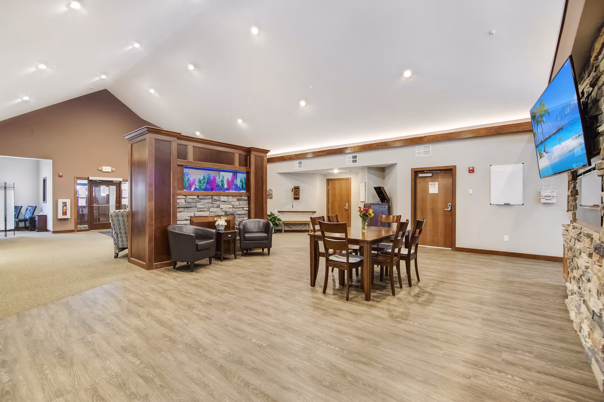 A spacious senior living common area with a high vaulted ceiling and recessed lighting. The room features a wooden table with six chairs, two black leather armchairs with a small side table, and a large wooden partition with a built-in aquarium. A flat-screen TV is mounted on a stone wall to the right. The flooring is light wood, and there are wooden doors and neutral-colored walls in the background.