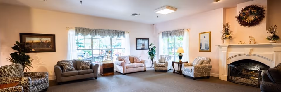 A spacious living room with multiple upholstered chairs and sofas arranged around the room. There are large windows with sheer curtains allowing natural light to fill the space. A fireplace with decorative items and a wreath above it is on the right side of the room. The walls are light-colored, and there are framed pictures and plants adding to the cozy atmosphere.