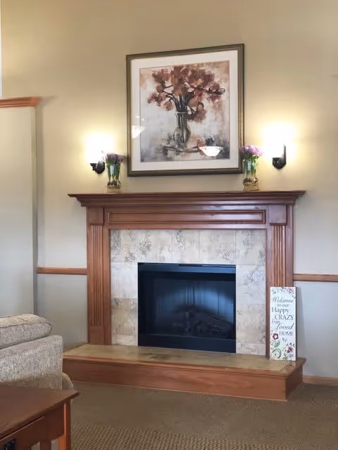 Interior living area showing a decorative wood-mantel fireplace with tile surround, a floral painting above, wall sconces, and part of a sofa.