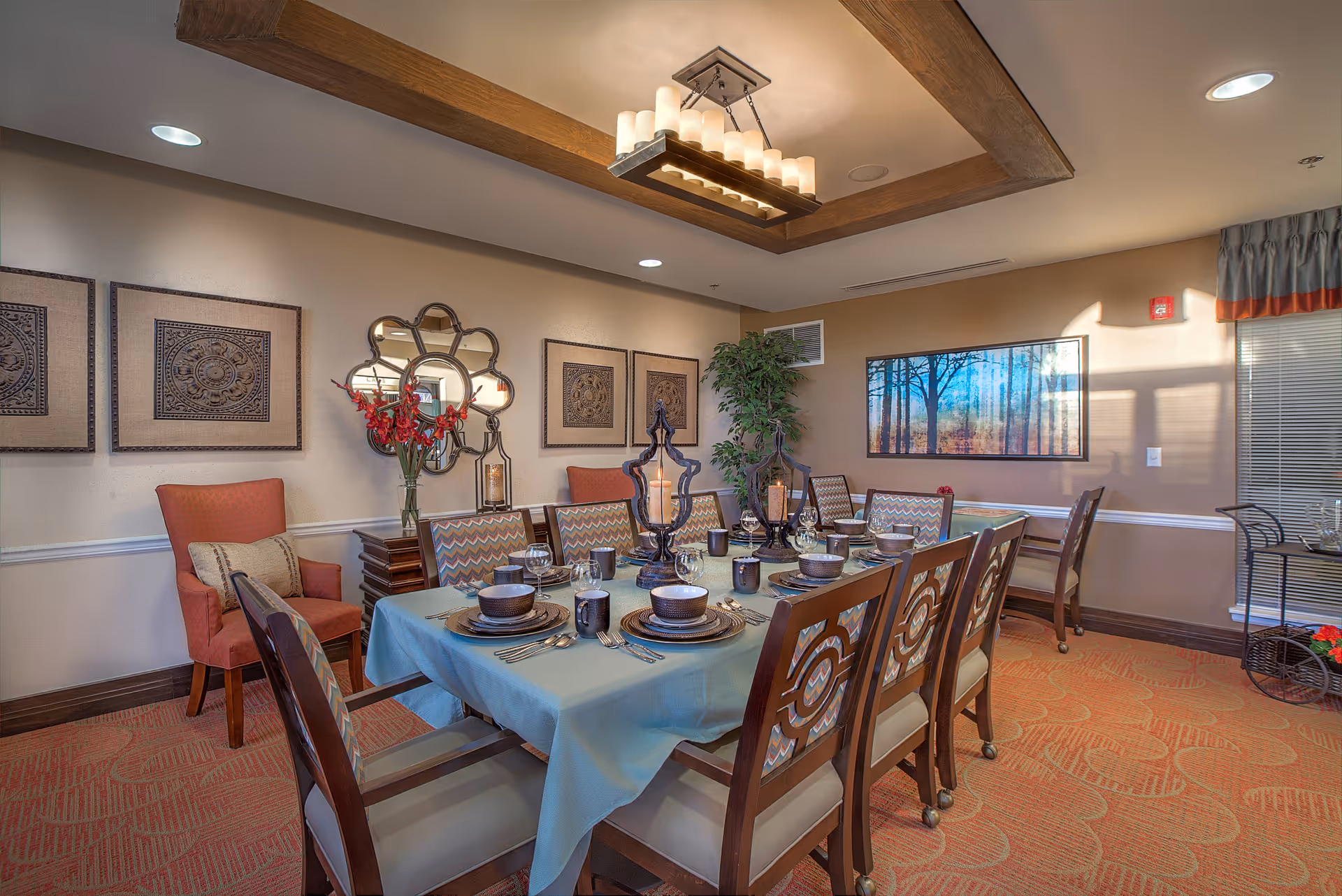 A well-decorated dining room with a rectangular table covered with a light blue tablecloth, set with plates, bowls, cups, and silverware. The table is surrounded by wooden chairs with patterned cushions. The room features warm lighting from a modern chandelier, wall art including a large framed picture of trees, decorative mirrors, and a potted plant in the corner. The carpet has a floral pattern, and there is an orange armchair with a cushion against the wall.