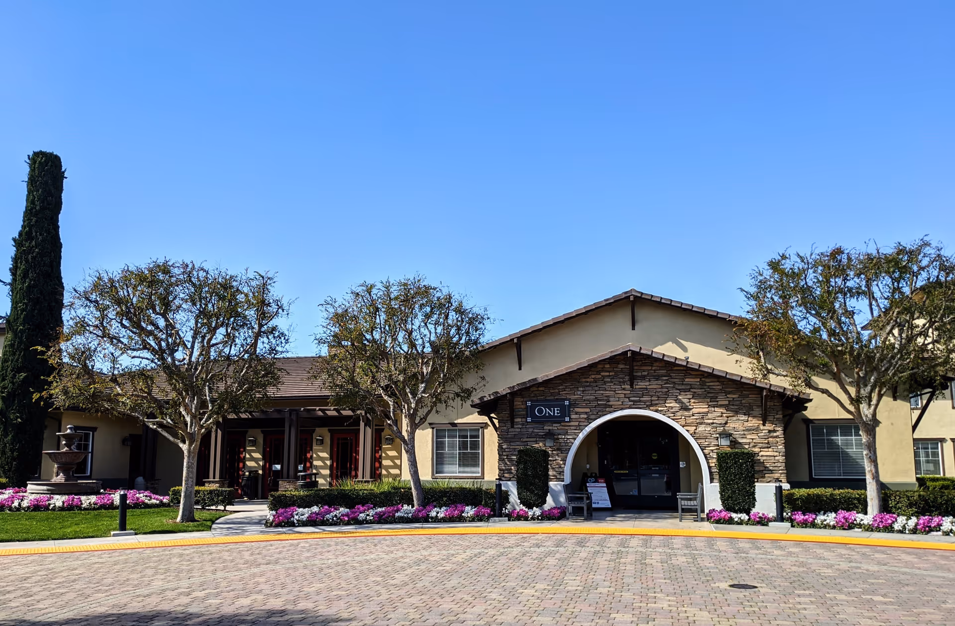 Front exterior view of Woodbridge Terrace facility building with a stone and stucco facade, an arched entrance, neatly trimmed trees, colorful flower beds, and a clear blue sky.
