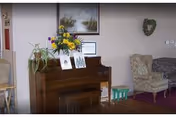 Interior common room with an upright piano topped with framed photos and flowers, upholstered armchairs, a wall painting, and a heart-shaped wreath.