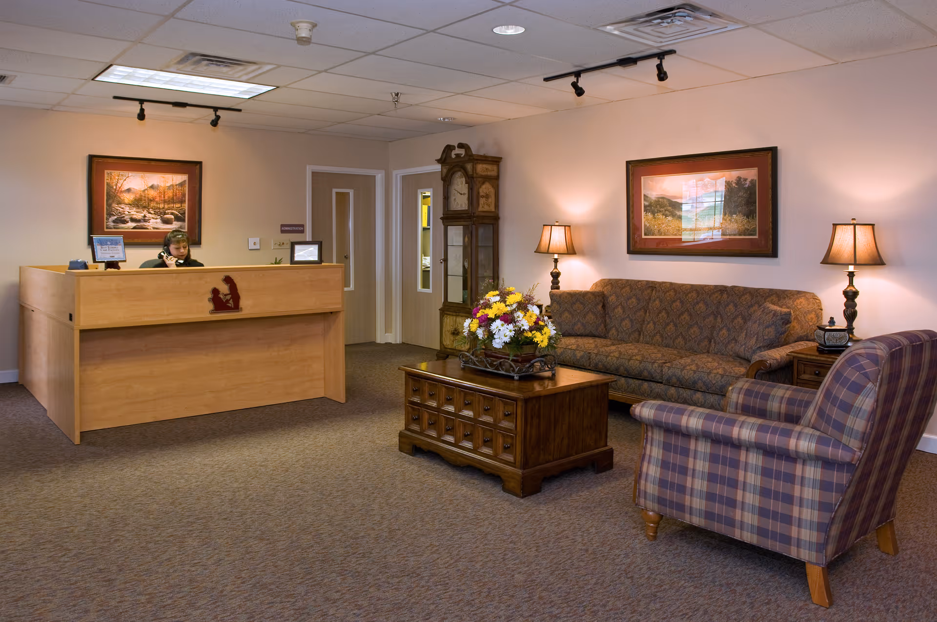 Reception/lounge area with a wooden front desk, a sofa and armchair around a coffee table topped with flowers, and lamps and framed art on the walls.