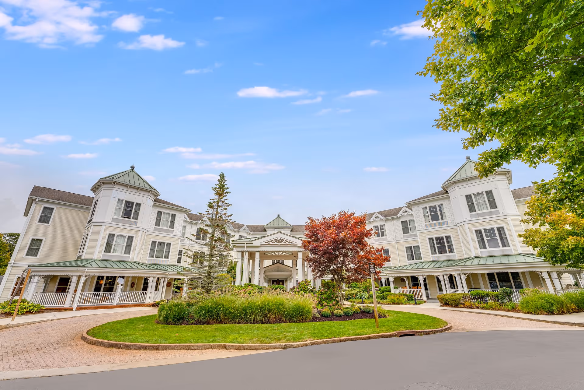 Front exterior view of a large, elegant senior living facility building with white siding, green roofs, and multiple windows. The entrance is covered with a white portico, and there is a circular driveway with landscaped greenery and trees, including one with red leaves, under a partly cloudy blue sky.