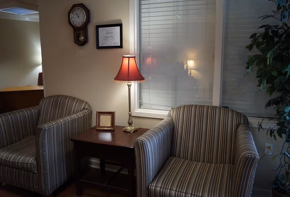 A cozy seating area with two striped armchairs separated by a wooden side table holding a lamp with a red shade and a small empty picture frame. On the wall above the table, there is a clock and a framed certificate. A large window with closed blinds is behind the table and chairs, and a leafy green plant is partially visible on the right side.