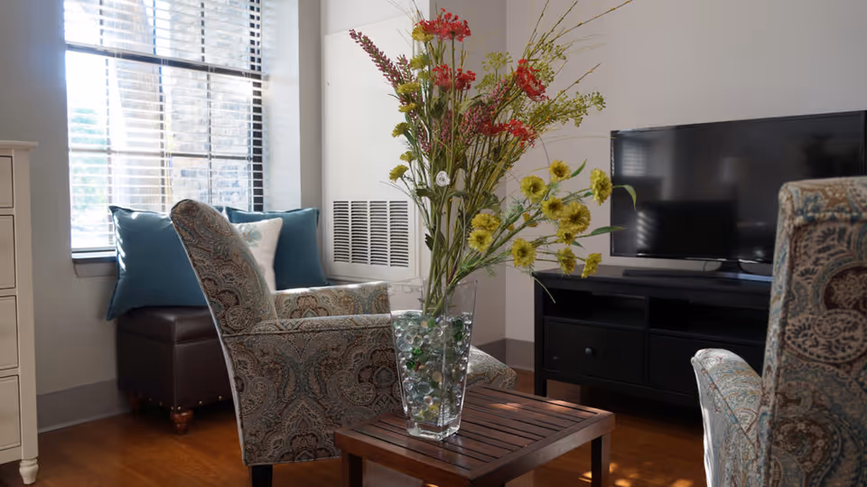 A cozy living room area with two patterned armchairs facing a wooden coffee table that holds a glass vase filled with decorative marbles and tall flowers. Behind one chair is a window with blinds and blue cushions on a dark ottoman. A flat-screen TV sits on a black media console against the wall.