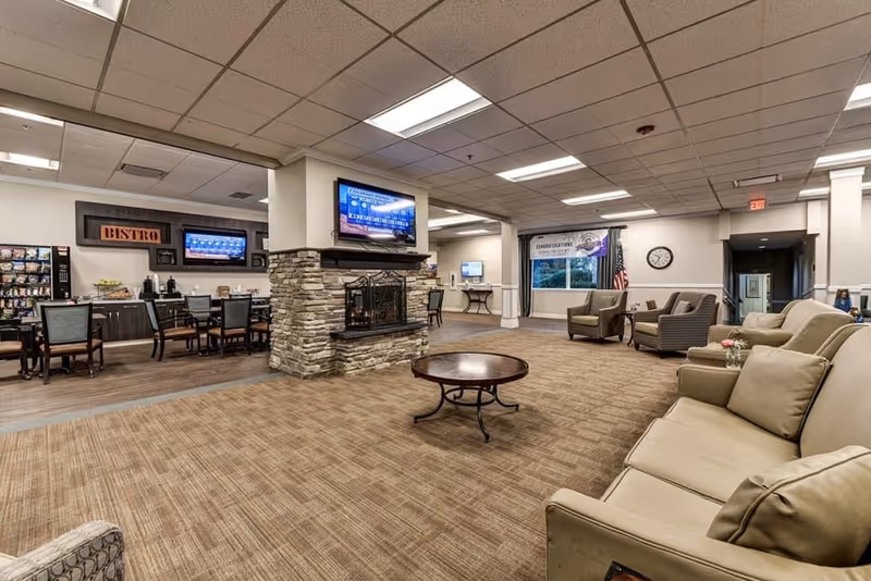 A spacious common area in a senior living facility featuring a stone fireplace with a mounted TV above it, comfortable seating including sofas and armchairs, a round coffee table, and a dining area with tables and chairs labeled 'Bistro'. There is a vending machine, a clock on the wall, and a banner near a window with an American flag beside it.