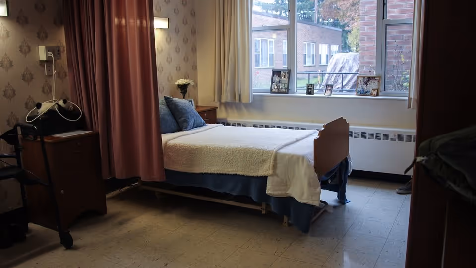 A small bedroom in a senior living facility with a single bed covered with a white blanket and blue pillows. There is a wooden nightstand with a vase of flowers and framed photos on the windowsill. A window shows an outside view of a brick building. A pink privacy curtain is partially drawn beside the bed, and a walker is positioned near the nightstand.