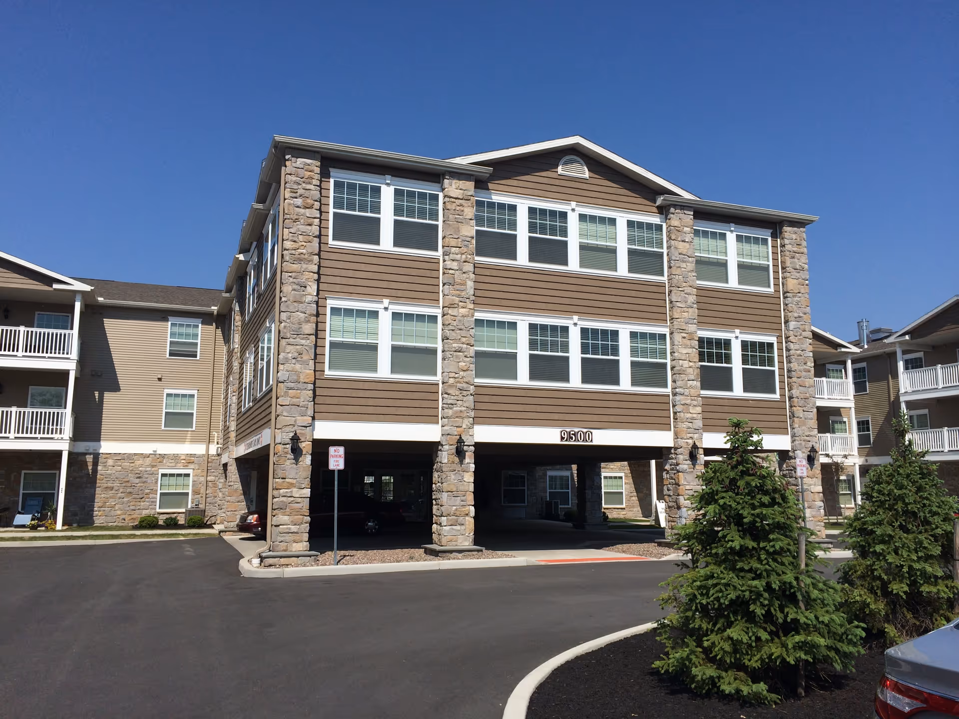 Exterior view of a multi-story senior living facility building with stone and wood siding, multiple windows, and a covered parking area underneath. The building number 9500 is visible above the parking entrance. There are small trees and landscaped areas in front of the building under a clear blue sky.