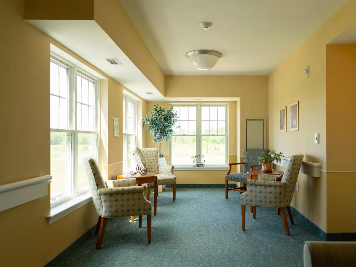 A bright sitting area in an assisted living facility with four upholstered chairs arranged around two small wooden tables. The room has large windows letting in natural light, beige walls, green carpet, and a potted plant near the window.