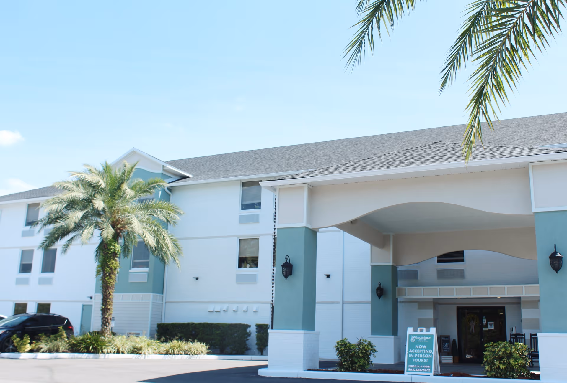 Front entrance of a two-story senior living building with a covered porte-cochere, palm tree, and an entrance sign.