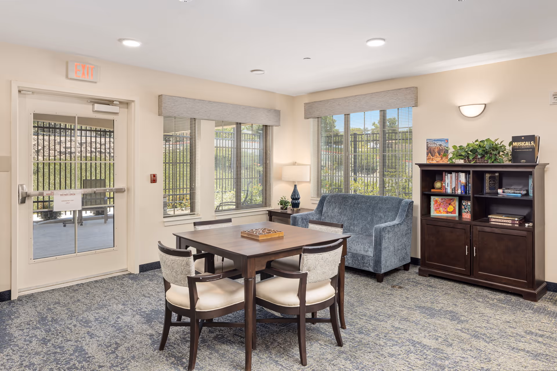 A cozy common area in a senior living facility featuring a wooden table with four upholstered chairs, a blue upholstered loveseat, a wooden bookshelf filled with books and decorative items, a table lamp, and large windows with blinds letting in natural light. An exit door with a sign is visible on the left side.