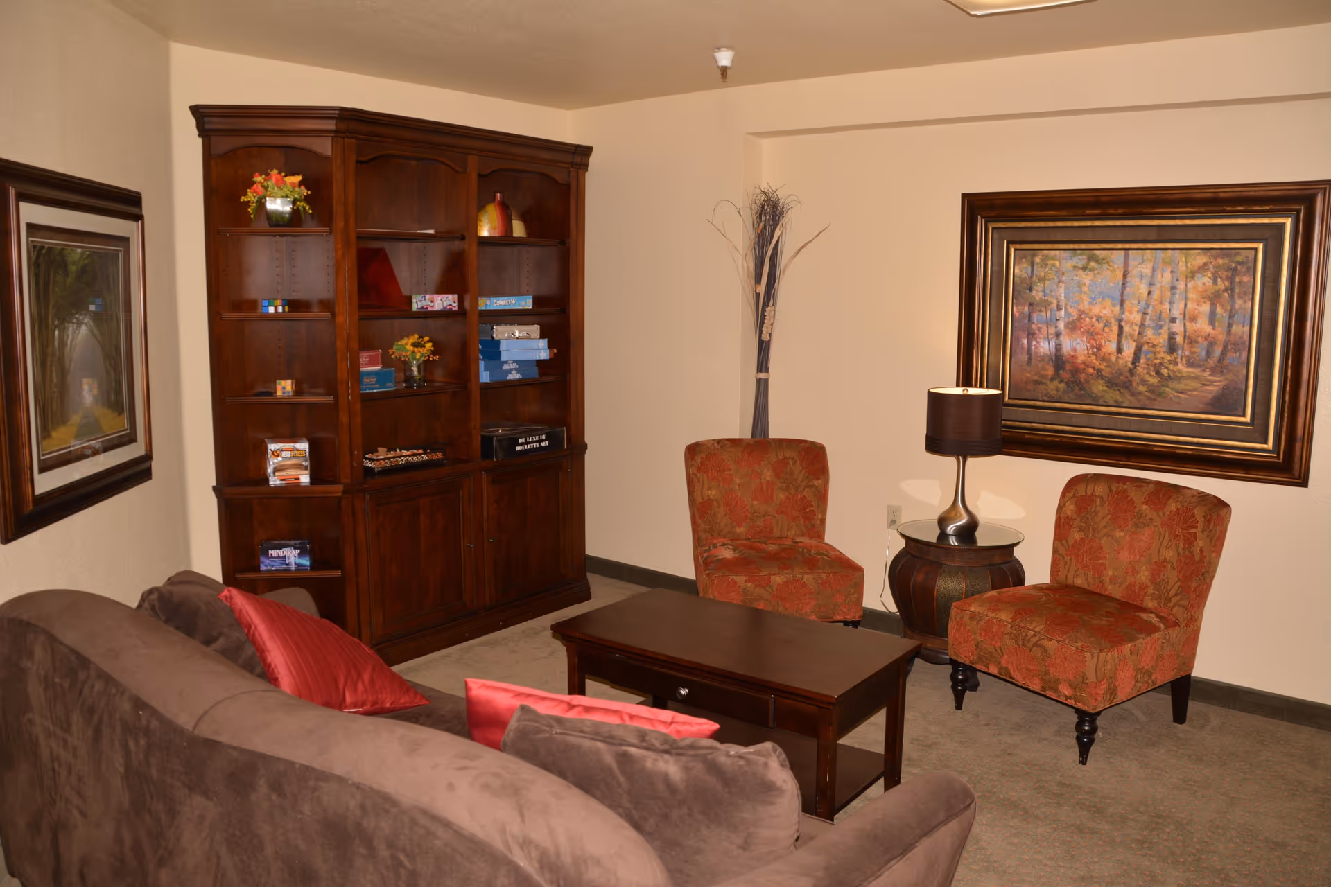 Cozy living room with a brown sofa, coffee table, two patterned chairs, a wooden bookshelf, and framed artwork on the walls.