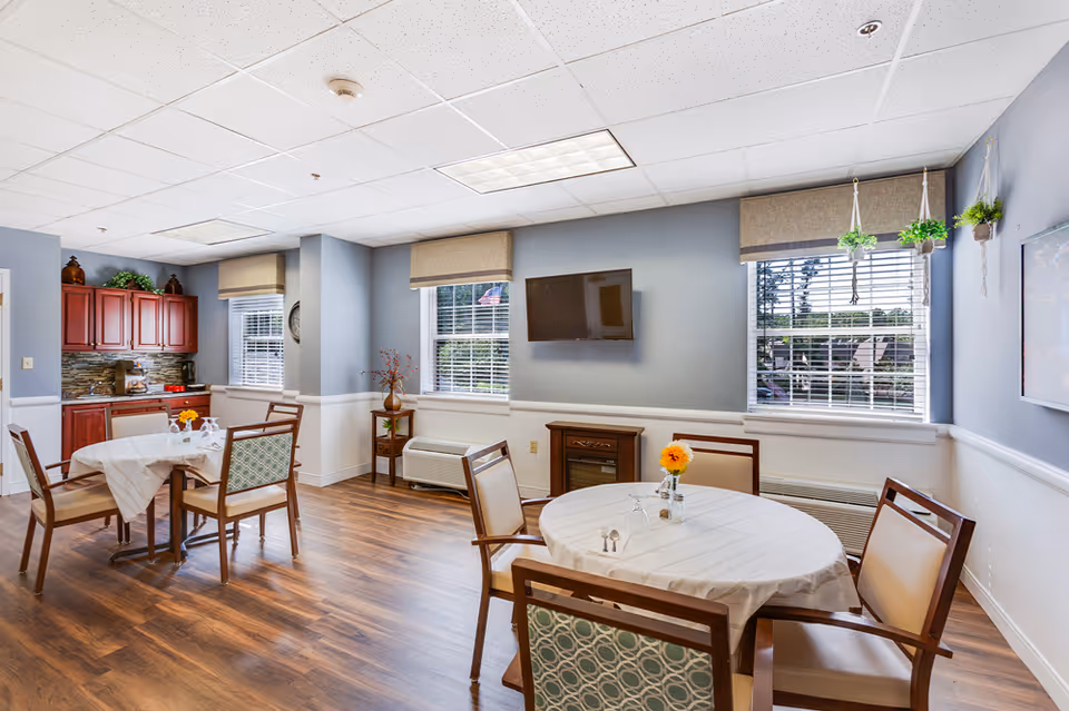 A bright dining area in a senior living facility with two round tables covered with white tablecloths, each surrounded by four chairs. The room has light blue walls, wood flooring, and large windows with beige blinds letting in natural light. A flat-screen TV is mounted on the wall, and there are hanging plants near one window. In the background, there is a small kitchen area with red cabinets and a stone backsplash.