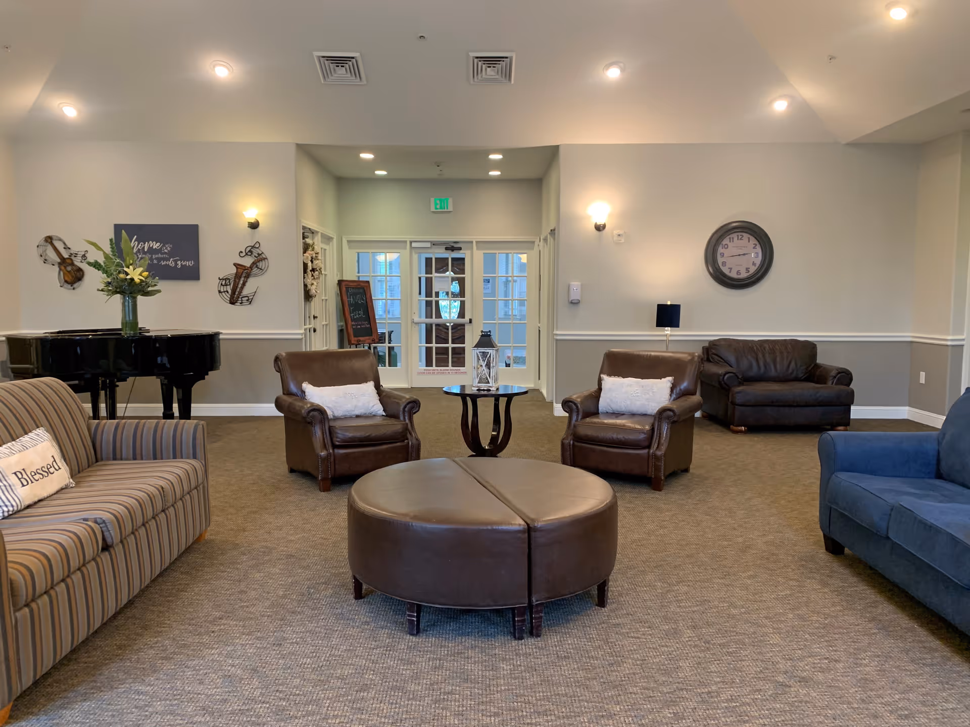 A cozy living room area in a senior care facility featuring a variety of seating options including a striped sofa with a 'Blessed' pillow, two brown leather armchairs with white pillows, a dark brown leather loveseat, and a blue sofa. In the center is a round, brown leather ottoman. The room has beige walls with white trim, a wall clock, wall sconces, a black grand piano with a vase of flowers, and decorative musical instrument wall art. Double glass doors lead to another area, and a small round table with a lantern is placed between the armchairs.