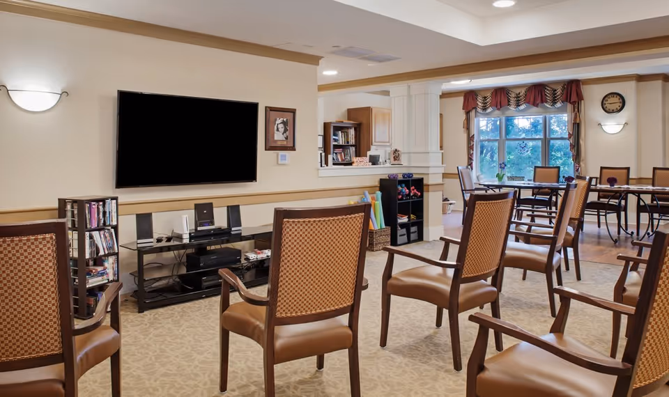 A senior living facility common area with several wooden chairs arranged facing a large flat-screen TV mounted on the wall. There is a small bookshelf with DVDs and books, a media console with electronic devices, and a window with decorative curtains letting in natural light. Tables and chairs are visible in the background near the window.