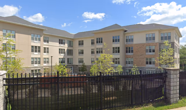 Exterior view of a multi-story senior living facility building with beige and brick facade, surrounded by a black metal fence and small trees under a partly cloudy blue sky.