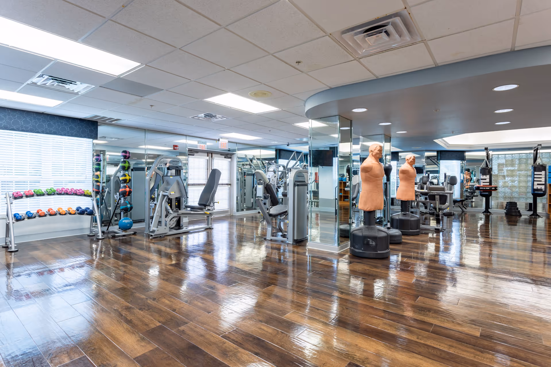 Bright fitness room with weight machines, colorful dumbbells, punching bags and freestanding dummy bags on polished wood floors and mirrored walls.