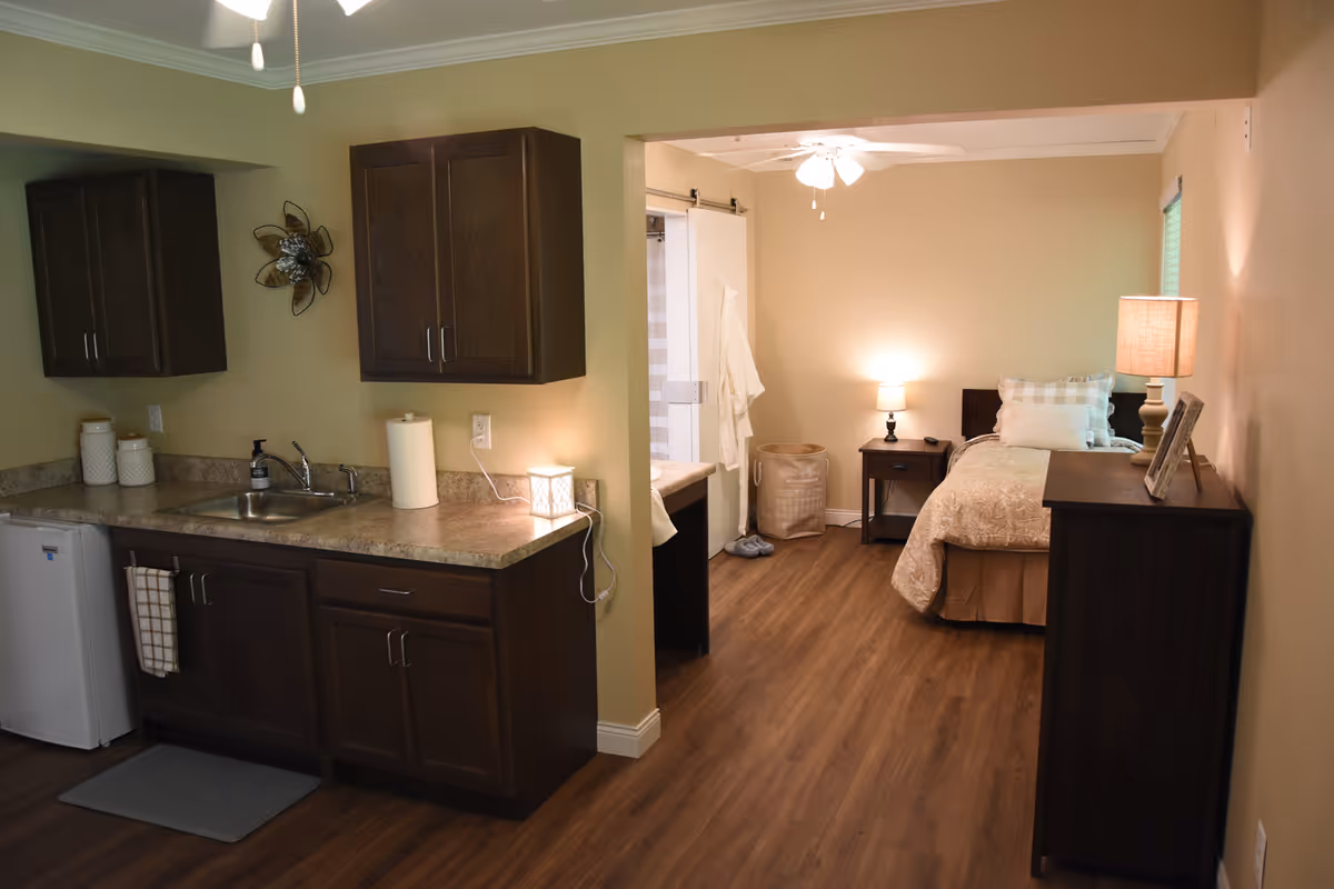 Interior view of a senior living facility room at Corridor Crossing Place showing a small kitchenette with dark wood cabinets, a sink, and a mini refrigerator on the left. To the right, there is a cozy bedroom area with a single bed, nightstand with a lamp, dresser with a lamp and picture frame, and a laundry basket. The room has wood flooring and warm beige walls, with ceiling fans and soft lighting.