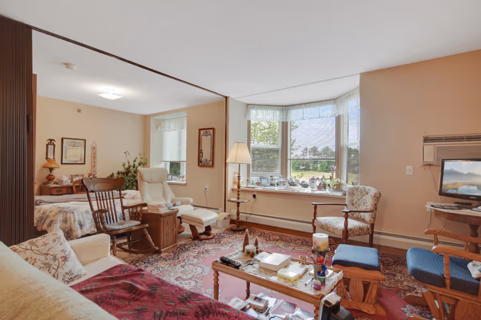A cozy living space in Elmhurst Commons featuring a floral-patterned sofa with a red throw, a wooden rocking chair, a white recliner with an ottoman, and several other upholstered chairs arranged around a wooden coffee table. The room has a large bay window with blinds and sheer curtains, letting in natural light and offering a view of greenery outside. The walls are painted beige, and the floor is covered with a patterned area rug. There are various decorative items on the coffee table and window sill, and a small TV is placed on a wooden stand in the corner.