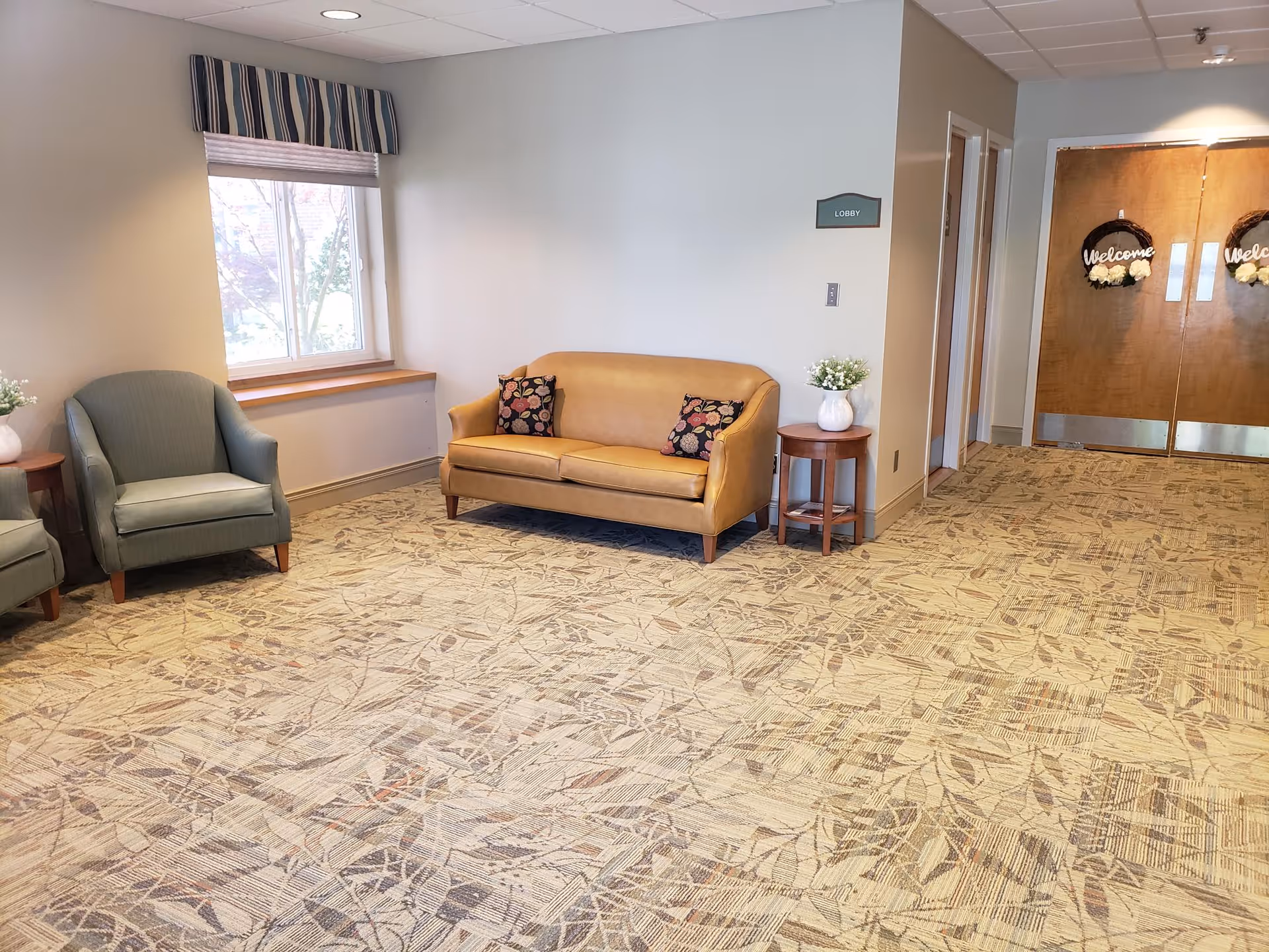 Lobby seating area with a tan sofa, two green armchairs, side tables with flowers, a window, and double doors in the background.