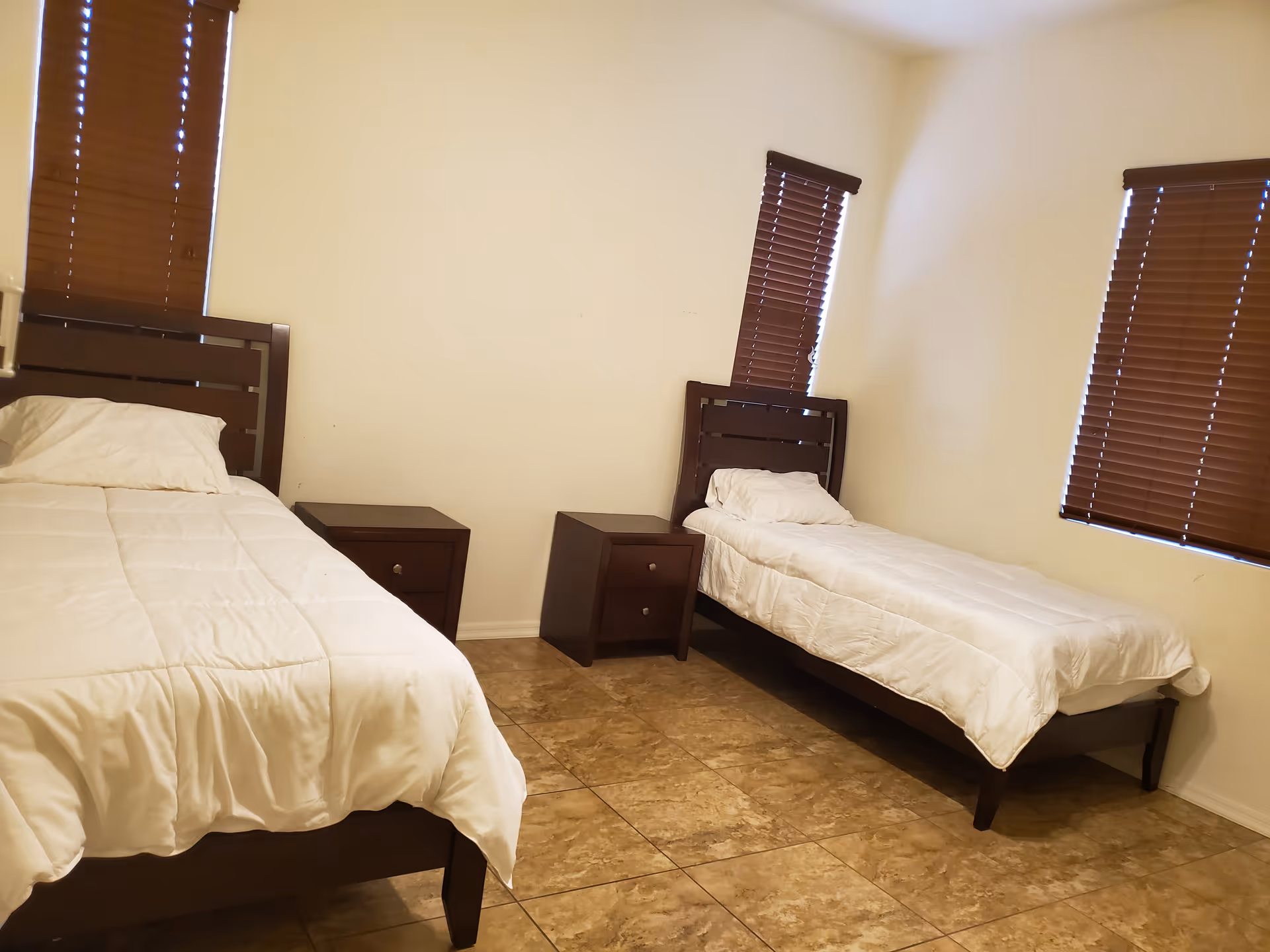 Sparsely furnished bedroom with two single beds, two nightstands, and brown window blinds.