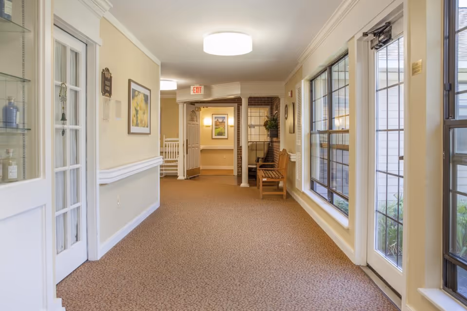 A well-lit hallway in a senior living facility with beige walls and carpeted floor. The hallway features large windows on the right side, a wooden bench, framed artwork on the walls, and a glass door on the left. At the end of the hallway, there is an open door leading to another room with additional seating and decor.