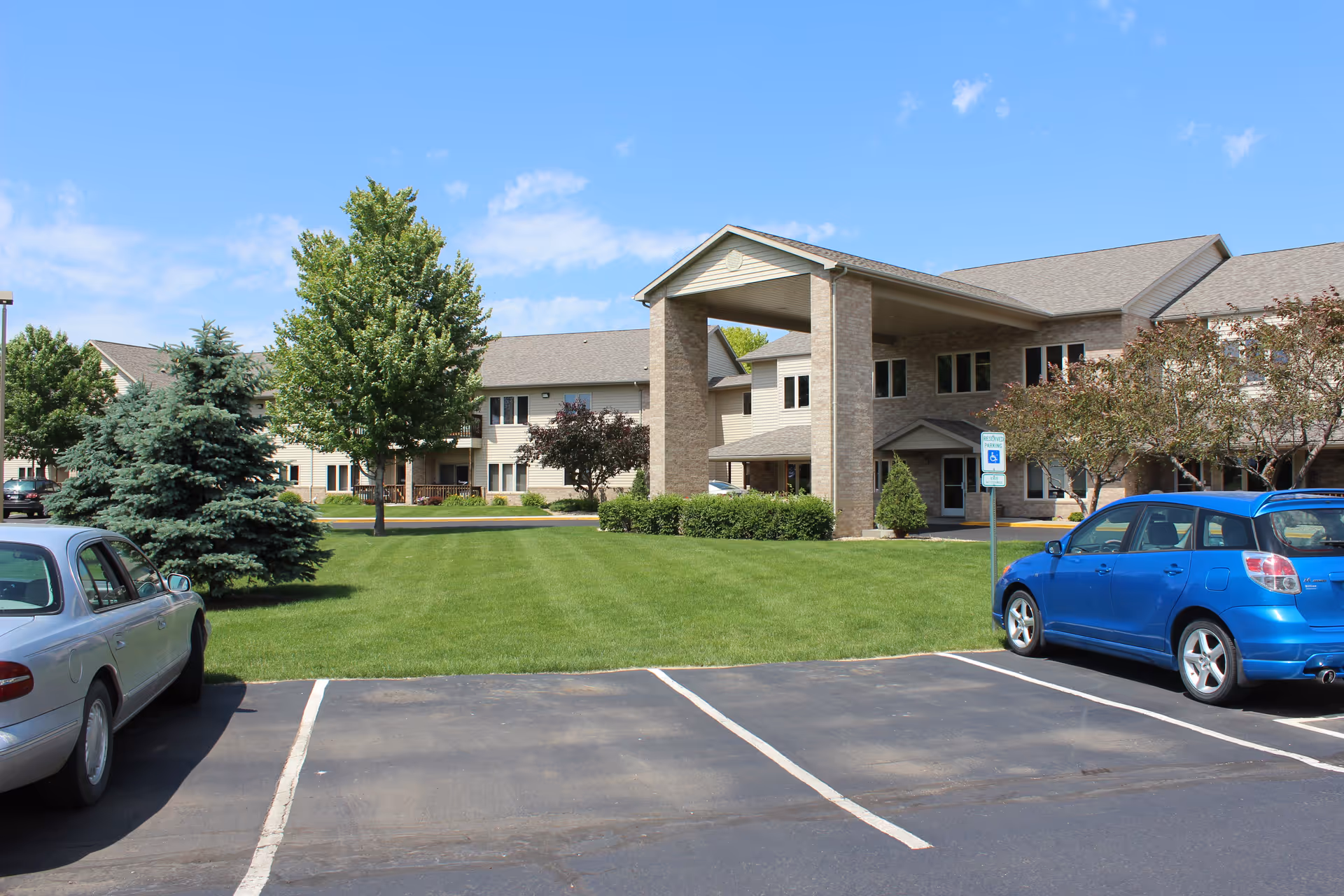 Exterior view of a senior living facility named Countryside Living with a well-maintained lawn, trees, and a parking lot with two cars parked. The building has a covered entrance and multiple windows under a clear blue sky.