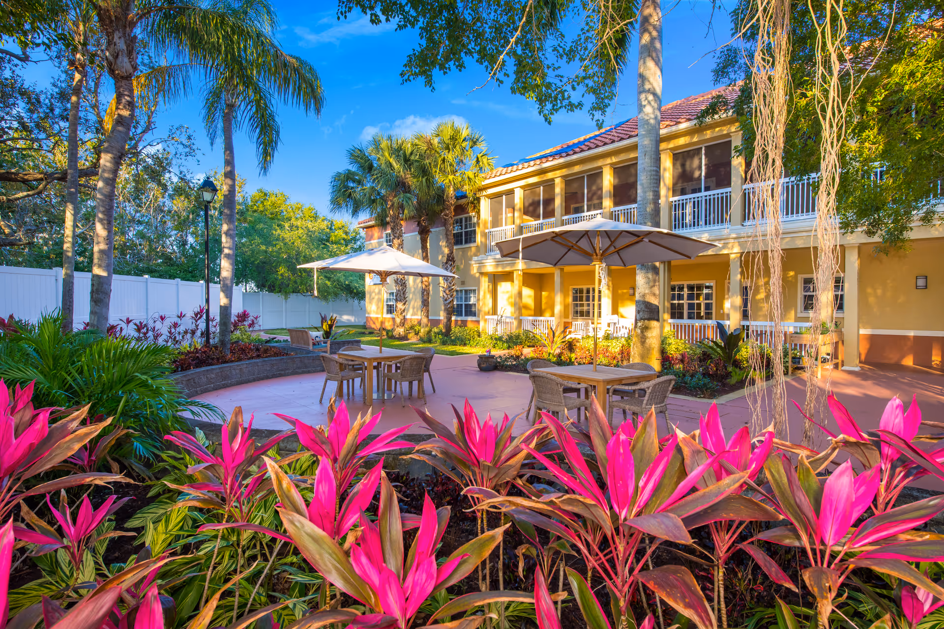 Sunny courtyard with patio tables and umbrellas surrounded by tropical plants and palm trees in front of a two-story senior living building.
