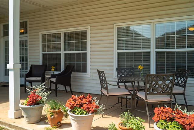 Outdoor patio area with a round metal table and four chairs, two additional chairs with a small table between them, and several potted plants with red and green foliage placed along the edge of the patio in front of a beige siding wall with two windows.