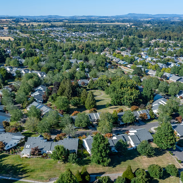 Aerial view of a tree-lined residential community with houses surrounding a central green park and distant hills.