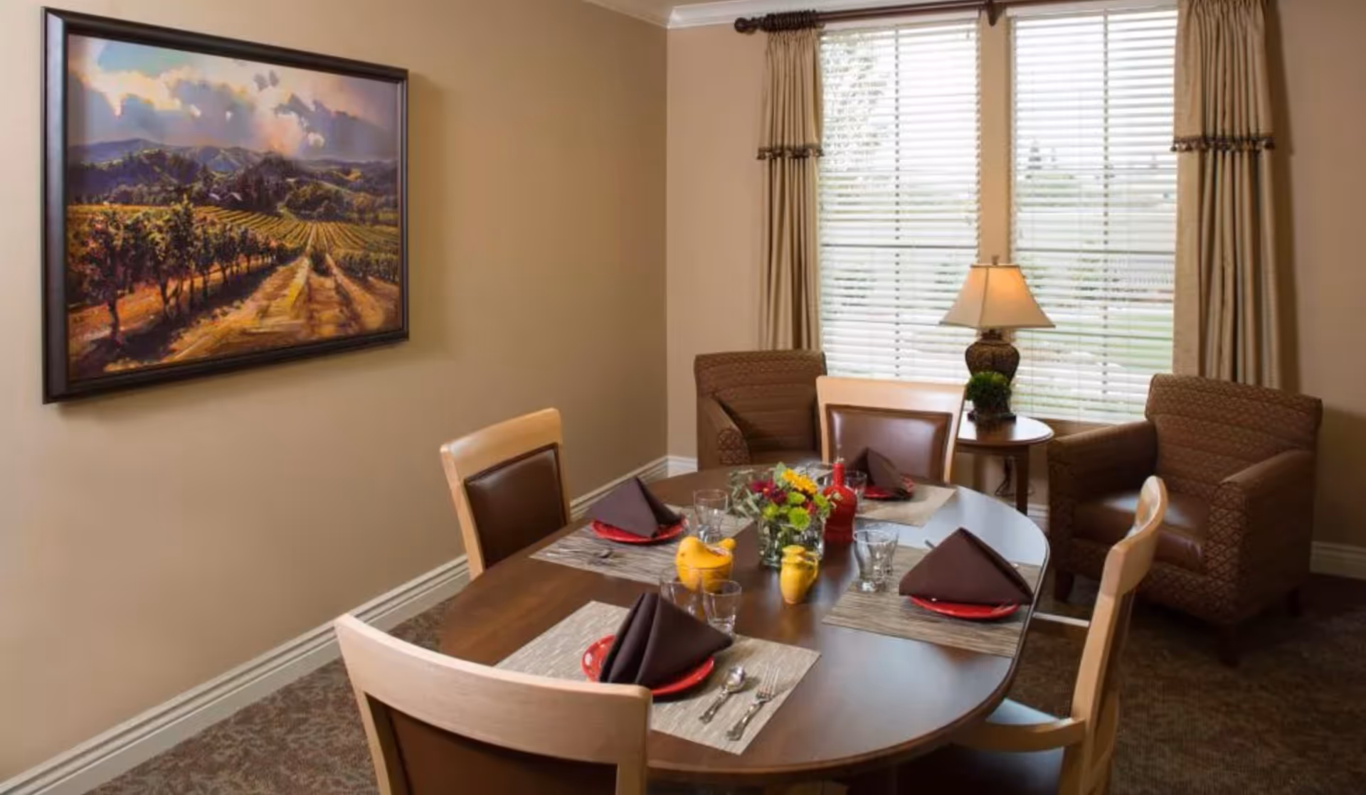 A dining room with a wooden oval table set for four people with brown napkins, red plates, glasses, and silverware. There is a small flower arrangement in the center of the table. The room has two upholstered armchairs near a window with blinds and beige curtains. A table lamp and a framed landscape painting are also visible.