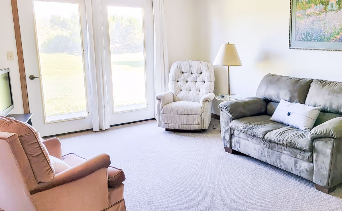 Sunlit living room with a couch, two armchairs, side table and lamp, and glass doors opening to a lawn.
