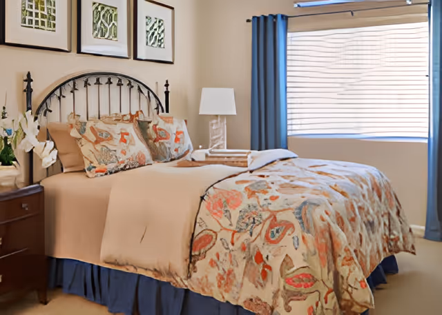 A neatly made bed with a floral patterned comforter and matching pillows in a bedroom. The bed has a black metal headboard. There is a wooden nightstand with a white orchid plant and a lamp with a white shade. Three framed artworks hang on the wall above the bed. A window with closed blinds and blue curtains is on the right side of the room.