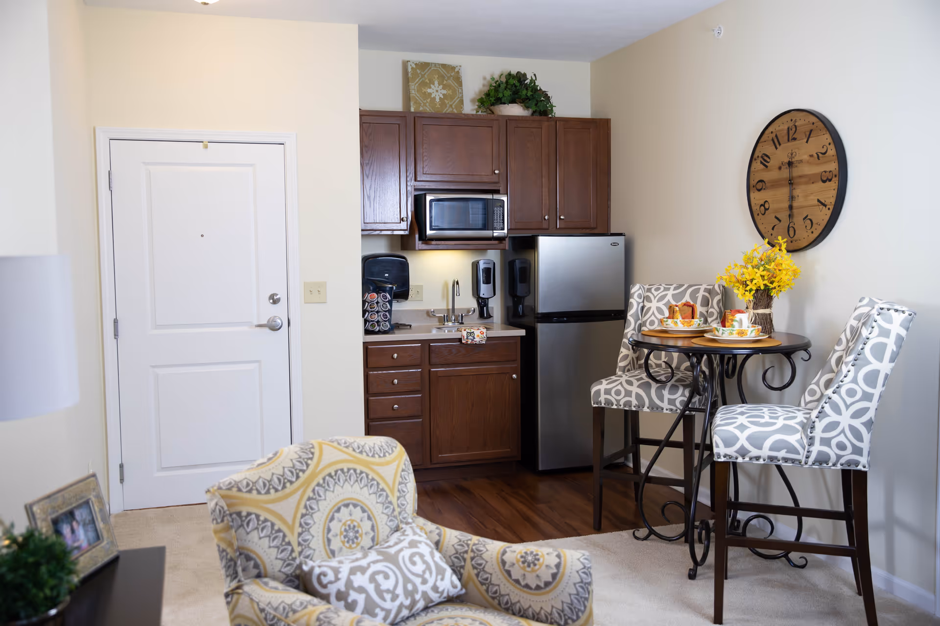 A cozy interior space featuring a small kitchenette with dark wooden cabinets, a microwave, a mini refrigerator, and a sink. Adjacent to the kitchenette is a small round table with two patterned high-back chairs, set with cups and plates, and a vase with yellow flowers. A large round wall clock hangs on the wall above the table. In the foreground, there is a patterned armchair and a white door on the left side of the image.