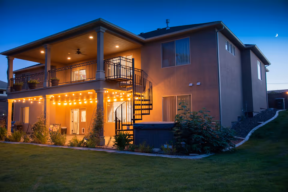 A two-story residential building at dusk with warm exterior lighting. The house features a covered balcony with a ceiling fan and potted plants, a spiral staircase leading from the balcony to the ground level, and string lights hanging under the balcony roof. The surrounding yard is grassy with some bushes and a paved pathway along the side of the house. The sky is clear with a crescent moon visible.