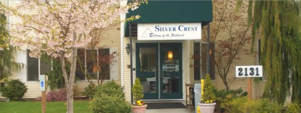 Entrance to Silver Crest, a senior living facility, featuring double glass doors under a green awning with the facility name and slogan. The entrance is surrounded by landscaped greenery, including trees and shrubs, with a sign displaying the number 2131.