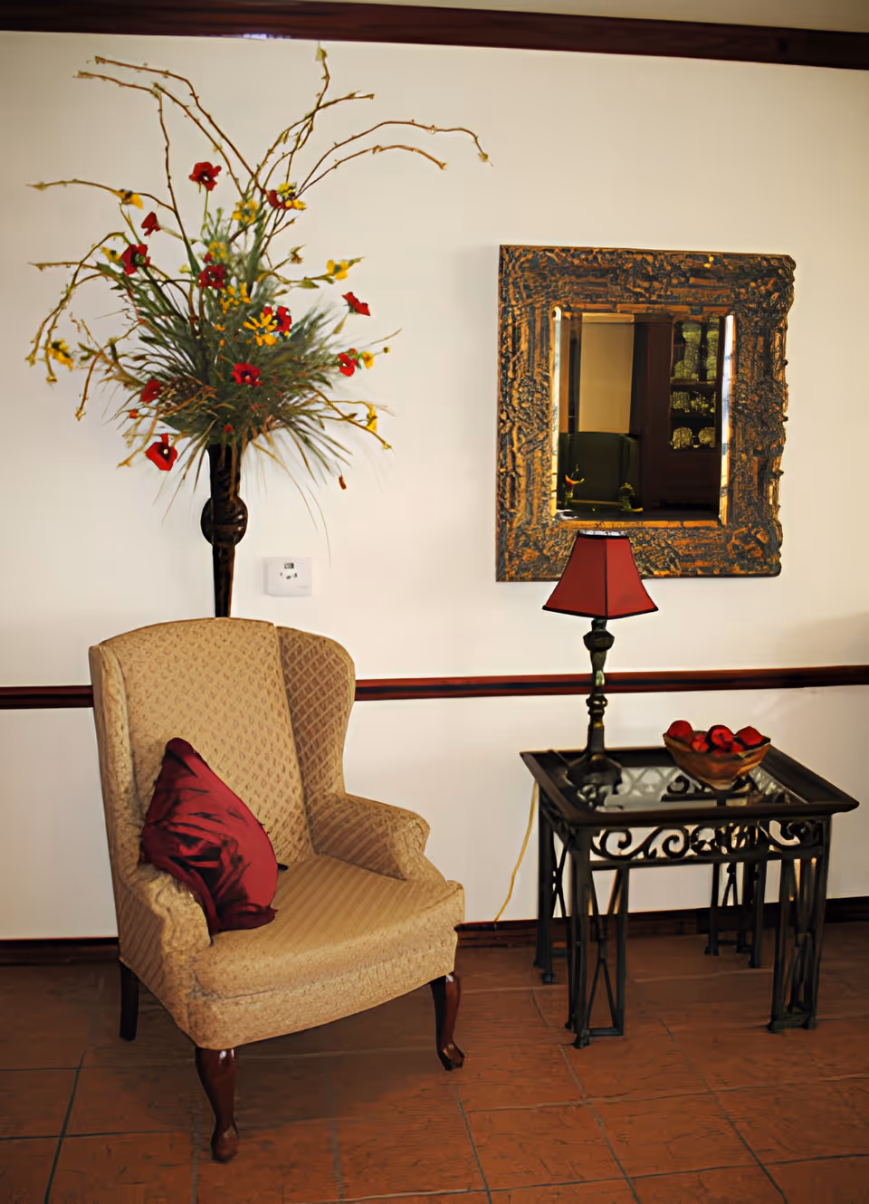 A cozy corner with a beige upholstered armchair featuring a red cushion, next to a black metal and glass side table holding a small red lampshade and a decorative bowl with red items. Above the table is a large ornate framed mirror reflecting part of the room. A tall floral arrangement with red and yellow flowers is mounted on the wall behind the chair.