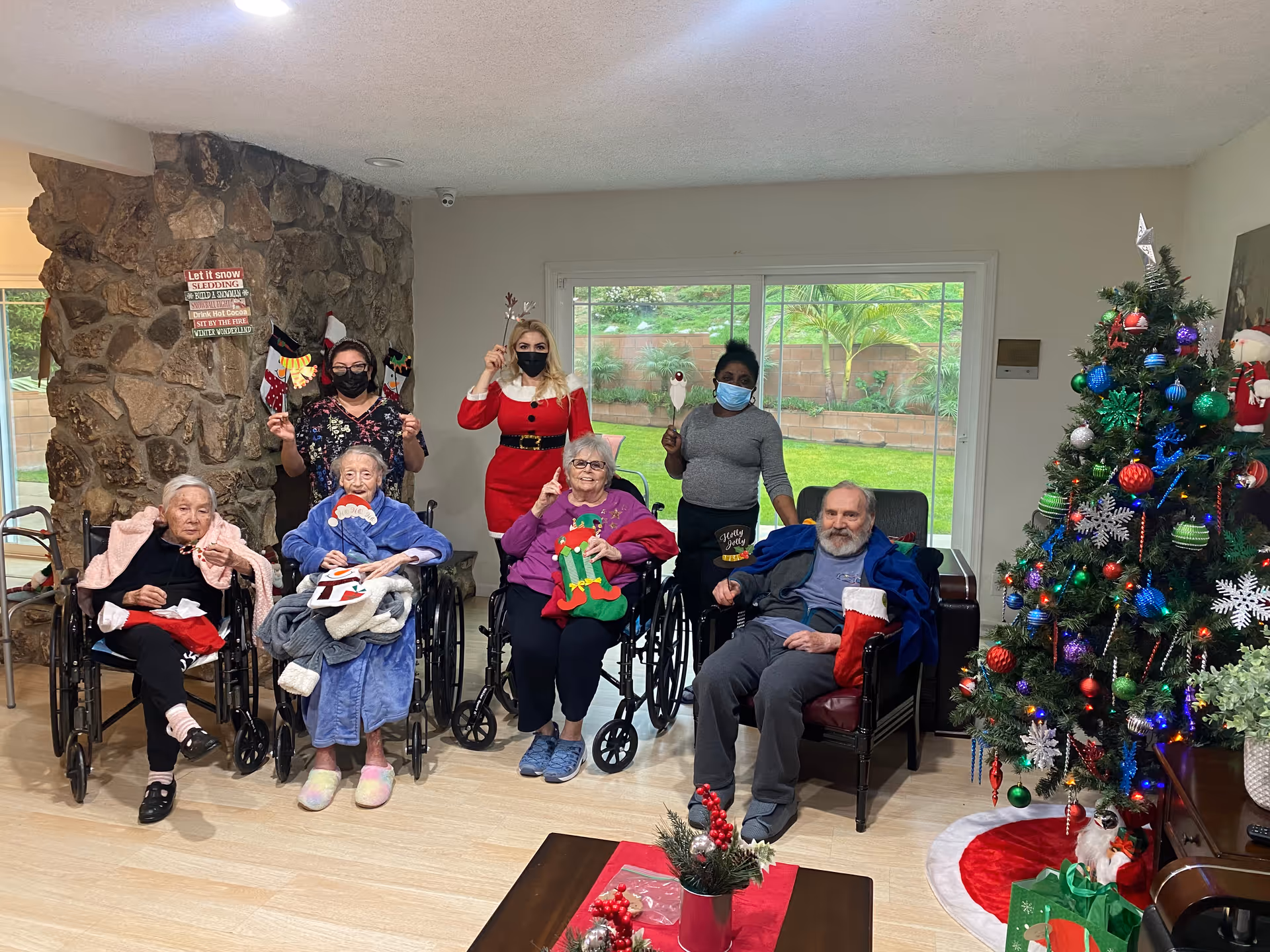 A group of elderly residents and two staff members posing in a festive living room decorated for Christmas. Four elderly people are seated in wheelchairs, and one elderly man is seated in a chair. Two staff members stand behind them, one dressed in a red holiday outfit and the other in casual clothes, both wearing face masks. The room has a stone wall with holiday signs, a large decorated Christmas tree with colorful ornaments and lights, and a sliding glass door showing a green garden outside.