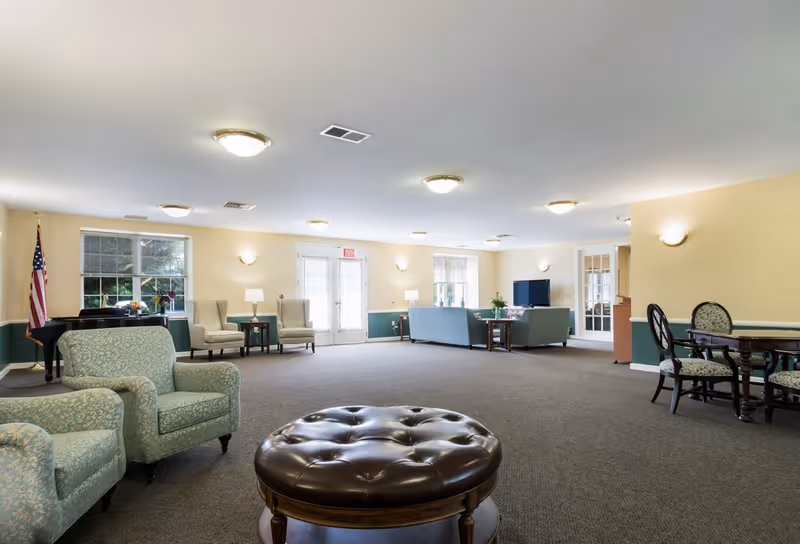 A spacious senior living facility common area with multiple seating arrangements including armchairs, sofas, and a round leather ottoman. The room has beige walls with green wainscoting, several windows letting in natural light, ceiling lights, a piano in the corner, and an American flag near the window.