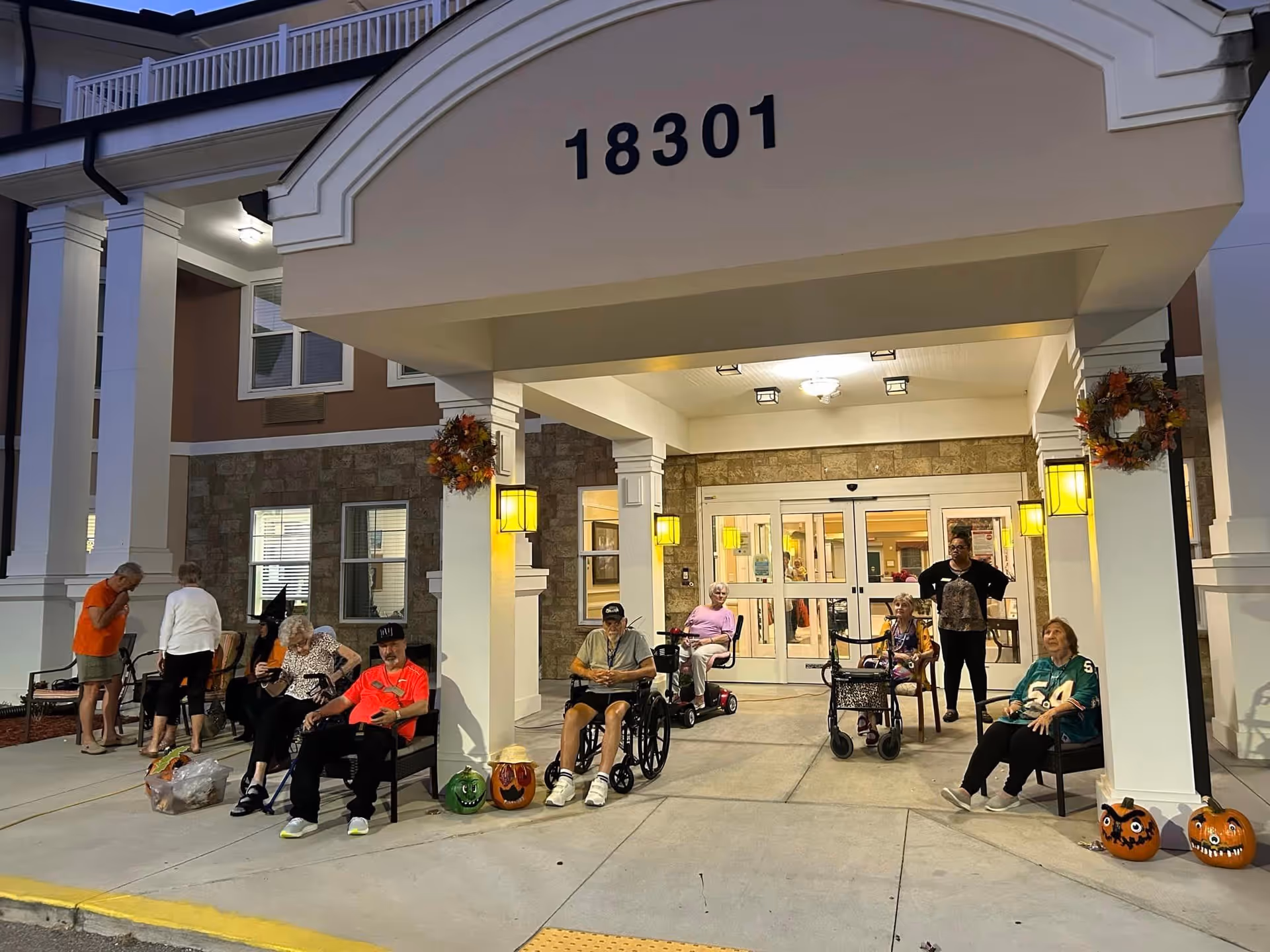Several elderly people sitting and standing outside the entrance of a senior living facility at dusk. The entrance has the number 18301 displayed above it, with fall-themed wreaths and decorated pumpkins placed around the area. Some individuals are seated in wheelchairs or on chairs, and a staff member stands nearby.