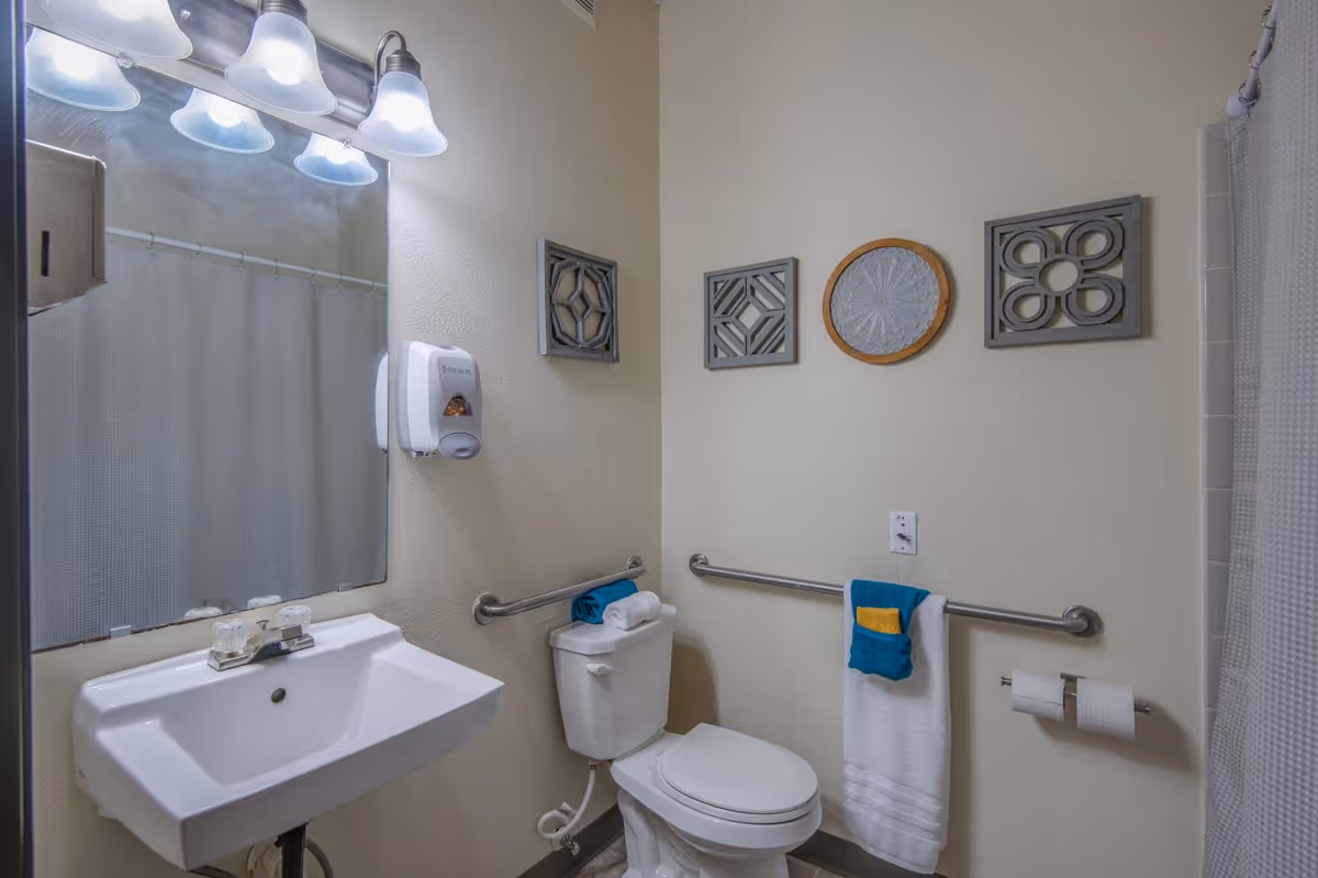 A clean bathroom with a white sink, mirror, and three light fixtures above it. There is a white toilet with a towel on top, a grab bar on the wall behind the toilet, and another grab bar with a towel hanging on it. The wall has three decorative pieces of art, and a shower curtain is visible on the right side.