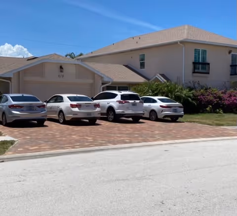 Four cars parked on a brick driveway in front of a two-story light-colored residential building and garage under a clear blue sky.