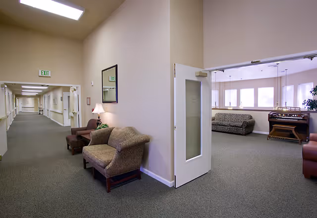 Interior view of a senior living facility hallway with beige walls and carpeted floors. There are armchairs and a small table with a lamp along the hallway wall. An open door leads to a common area with sofas, large windows, and a piano or organ.